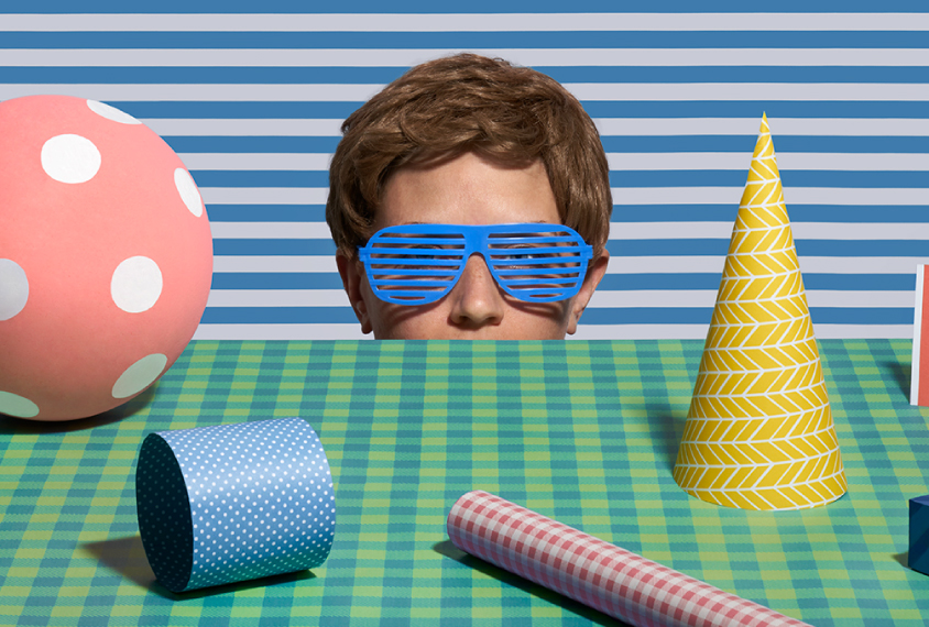 Humorous photograph shows a young man with plastic sunglasses on that match the patterns around him, surveys a tabletop scene of patterned objects.