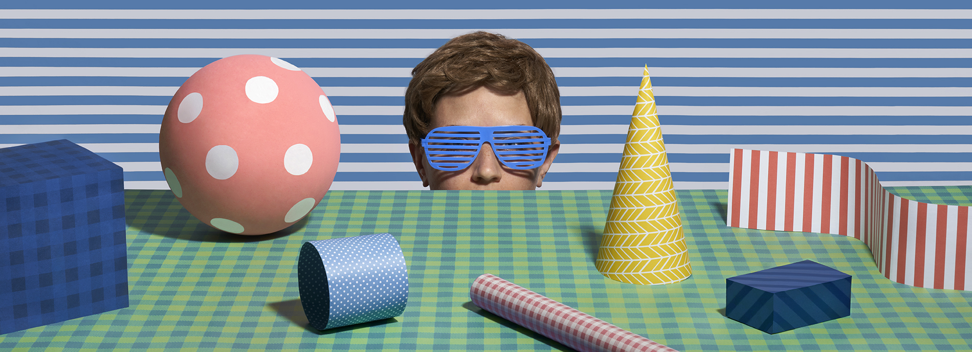 Humorous photograph shows a young man with plastic sunglasses on that match the patterns around him, surveys a tabletop scene of patterned objects.