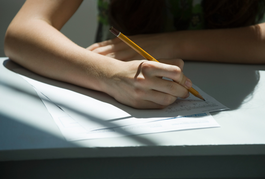 A student is seated at a table with a sheet of paper in front of him. They hold a pencil in this right hand and are filling out the sheet.