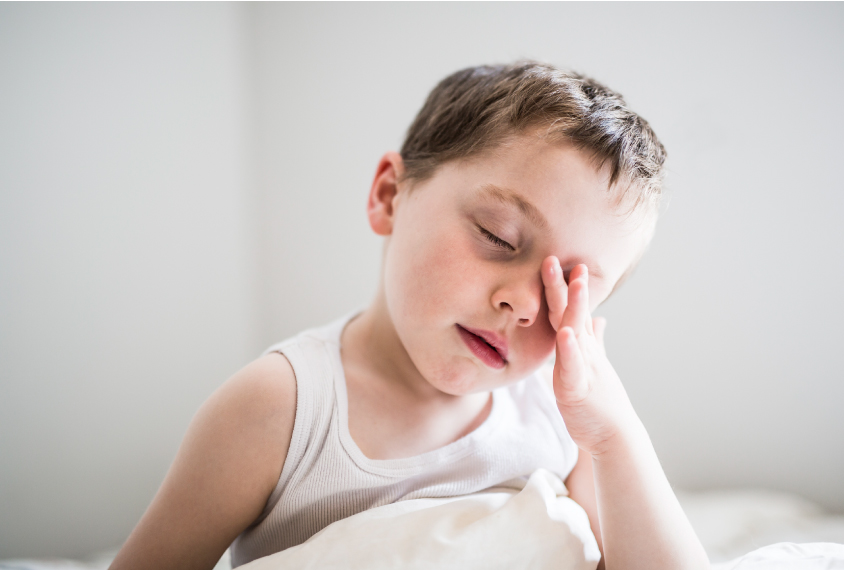 A young boy is sitting up in bed and wiping his left eye with his left hand.