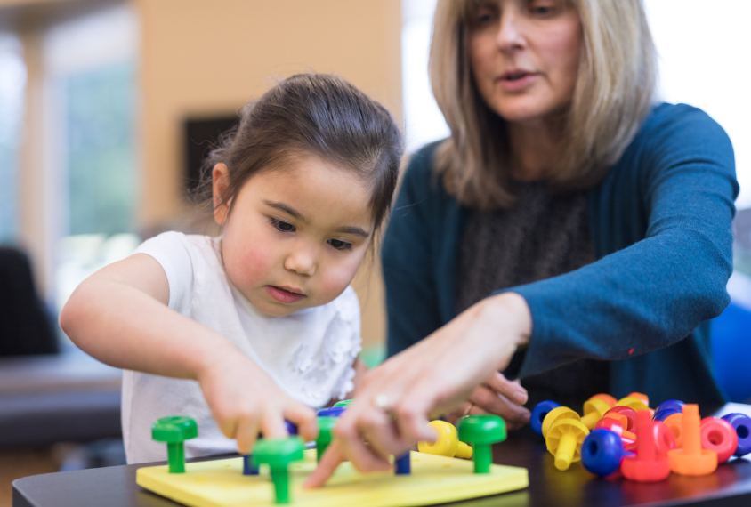 An occupational therapist works with a kindergarten-age girl on her coordination skills. She is showing her an exercise to practice putting pegs into a plastic board.