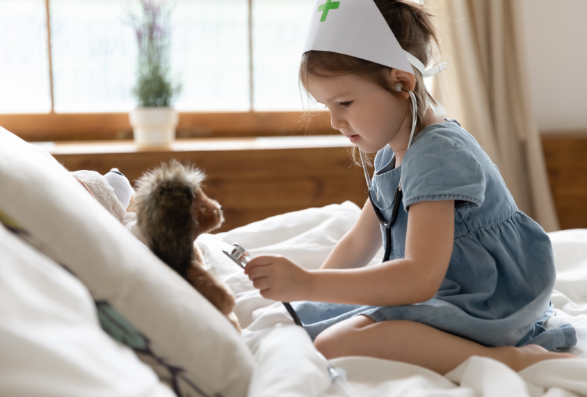 A young girl plays doctor on a bed with a stuffed animal.