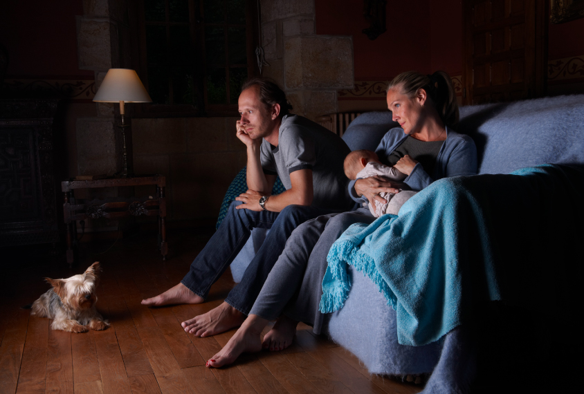 Parents sitting in the dark watching TV and nursing a baby.