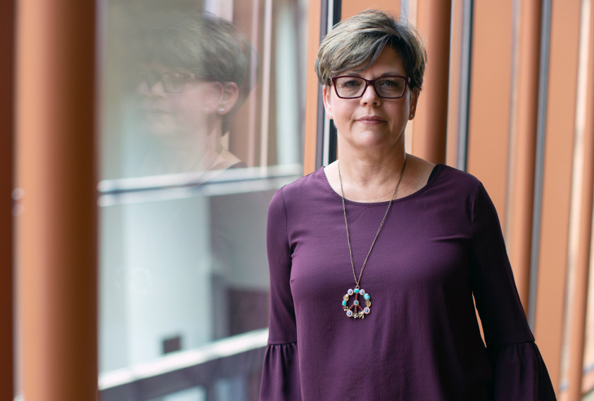 Photo: Social worker Laurie Ackles stands in front of a window. She's wearing a purple long-sleeved shirt, and she has short hair and glasses.