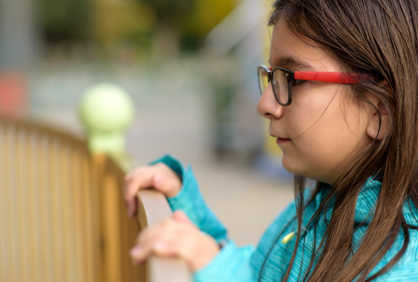 Elementary school age girl with glasses watches from outside a fence.