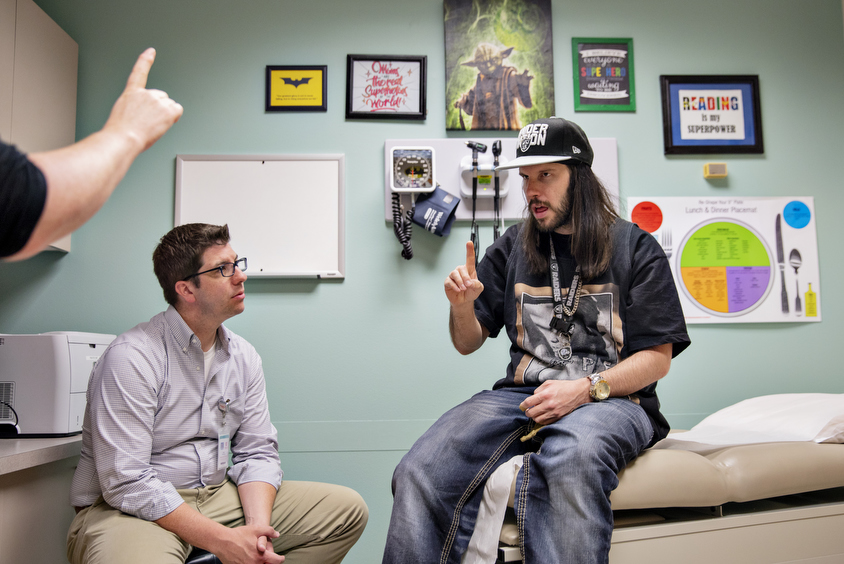 Photo: An autistic man sits on a doctor's bench, interacting with an interpreter out of frame. The doctor, sitting nearby, observes.