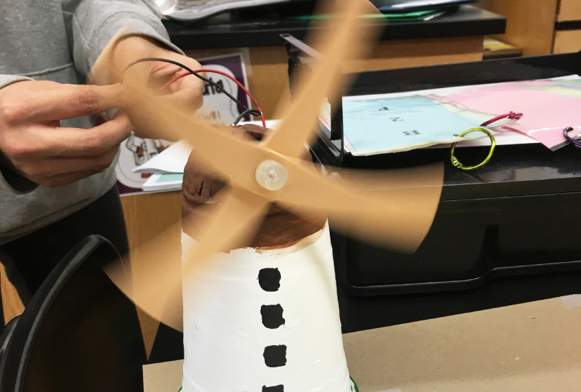 Classroom scene shows a kid powering up his handmade windmill model.