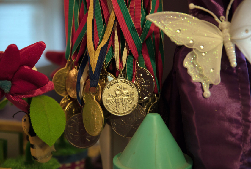 Photo: A close-up shot of hanging medals from Special Olympics competition. The metals are all shiny and gold-colored. Their ribbons are red, green, and yellow.
