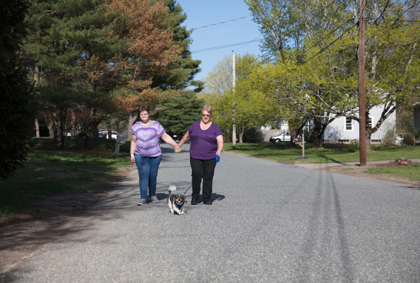 Photo: Becky, wearing a purple and white shirt and jeans, walks besides her mother, who is wearing a purple shirt and jeans. They're holding hands. The mother holds onto a leash for a small white and black dog. They're walking down a tree-lined neighborhood street.