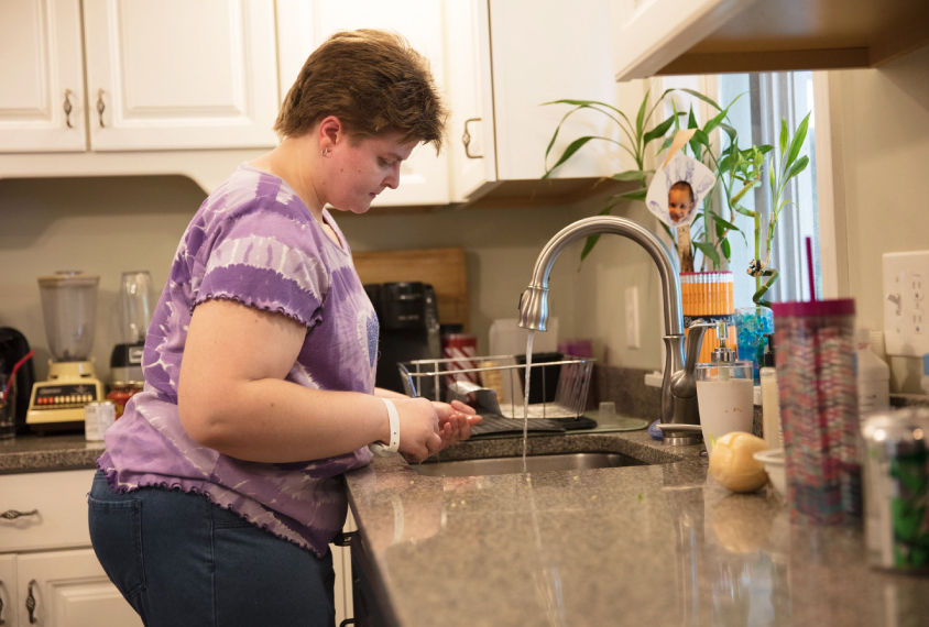 Photo: Becky Audette stands at the kitchen sink. She has short brown hair and is wearing a purple shirt and jeans. She is standing over the sink watching water pour from the faucet.