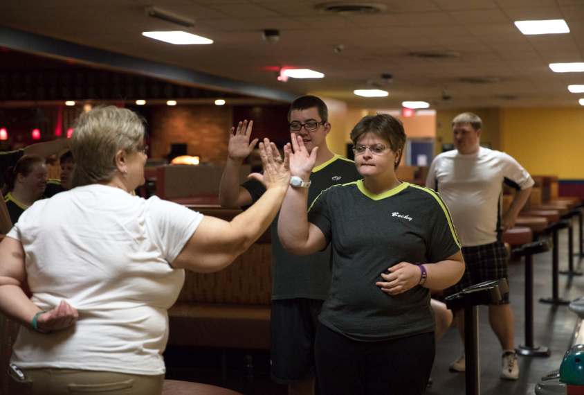 Photo: Becky Audette at a bowling alley. She's wearing a black and yellow bowling shirt. She high-fives a bowling teammate.