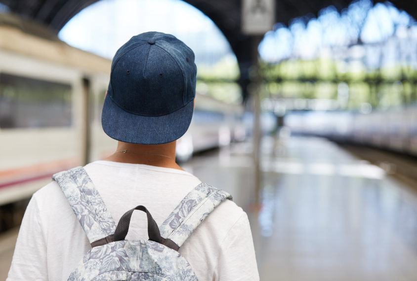 Boy in cap and backpack looks away from camera in train station.