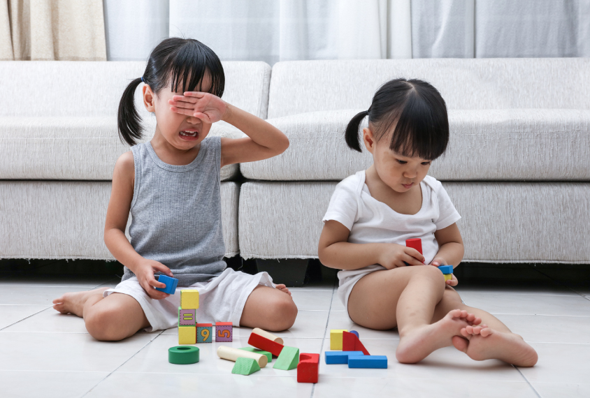 Two female children playing with blocks, one is crying, the other is facing away.