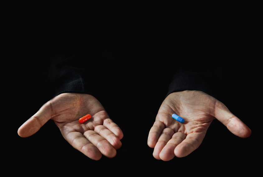 Two hands hold two pills, one red and one blue.