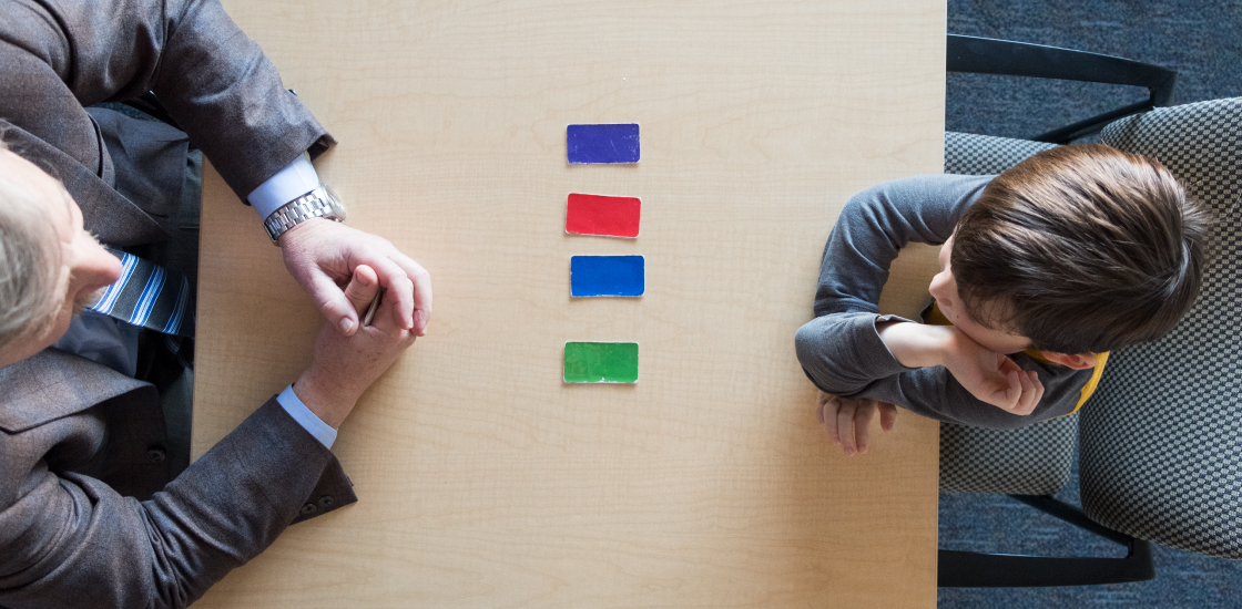 A doctor sits across from a child. On the desk between them are four cards, each of one color, to help test non-verbal intelligence.