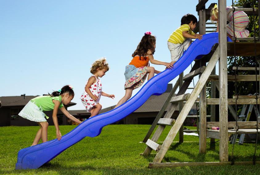 Group of young children climb up a blue slide outdoors on a sunny day.