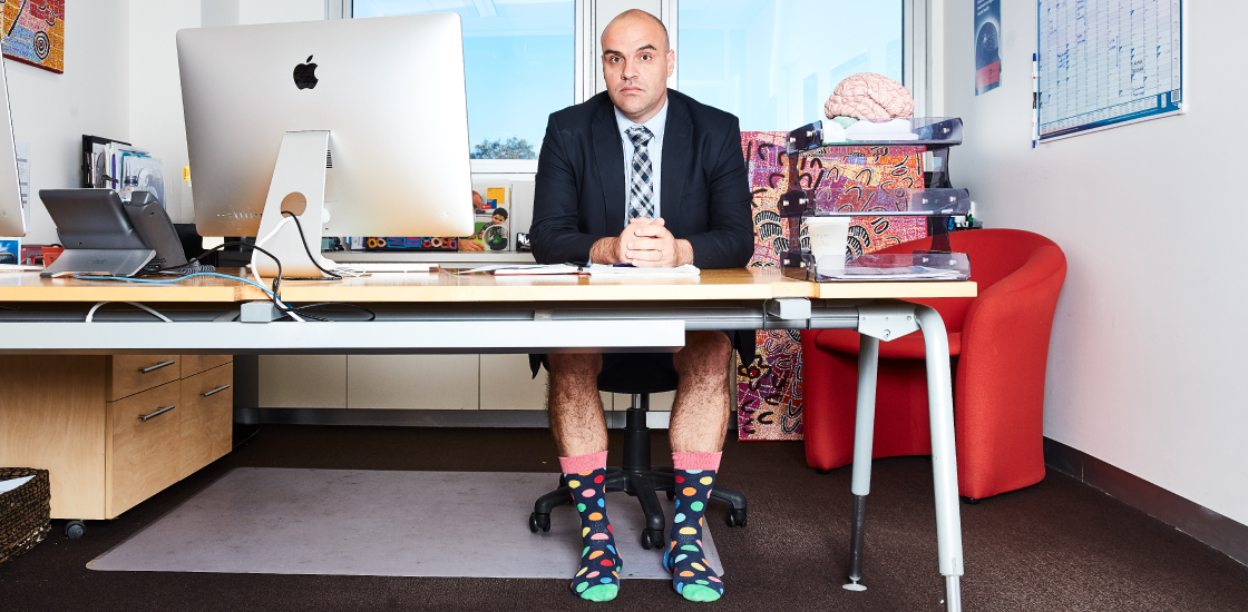 Goofy portrait of Adam Guastella at his desk in shorts and jacket with tie