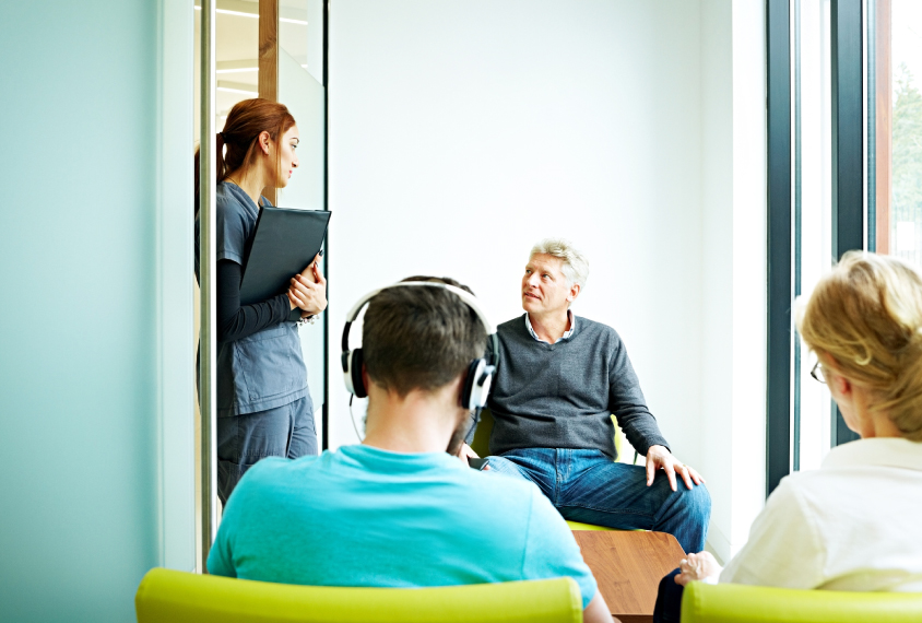 Adults in a waiting room, a man talks to a nurse or doctor in medical setting