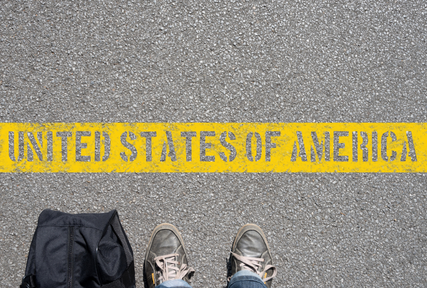 feet in worn sneakers stop in front of a yellow line on the pavement that reads: 'United States of America'