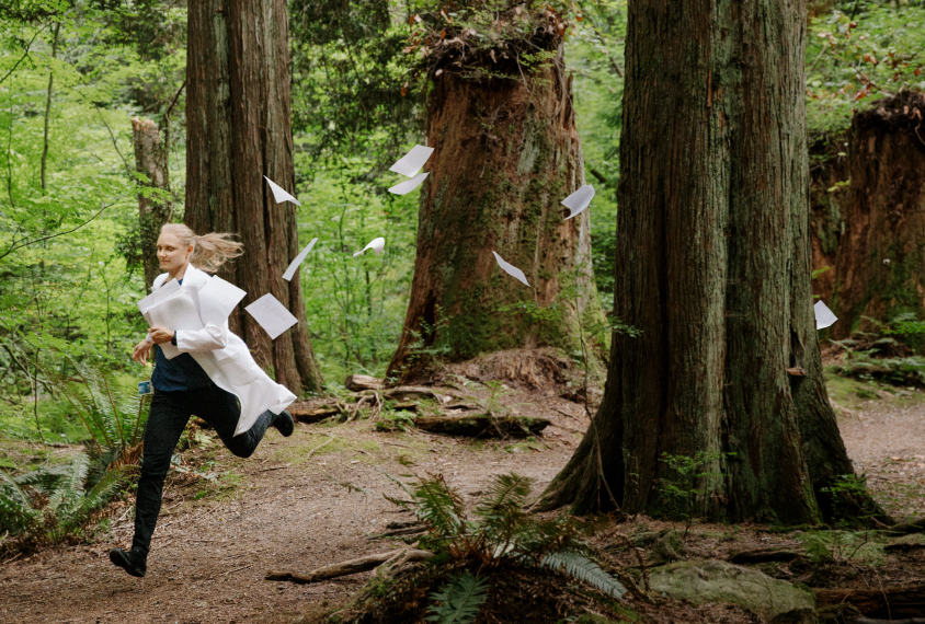 Scientist Annie Ciernia runs in the forest, wearing lab coat and carrying a stack of papers that are also flying along behind her