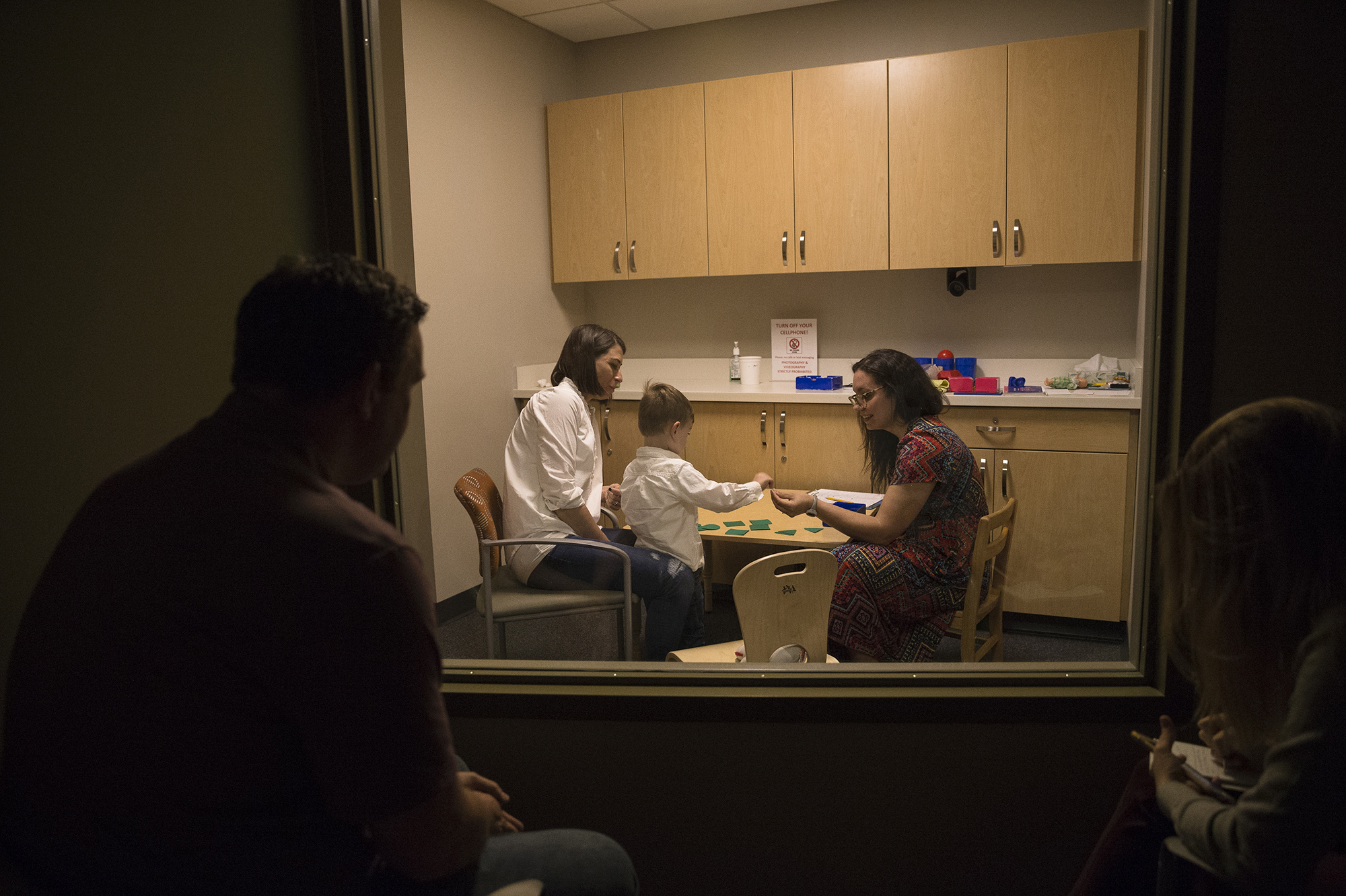Owen and his mother sit at a table with a clinician while she tests his ability to follow instructions. Owen's father watches through a window from another room.