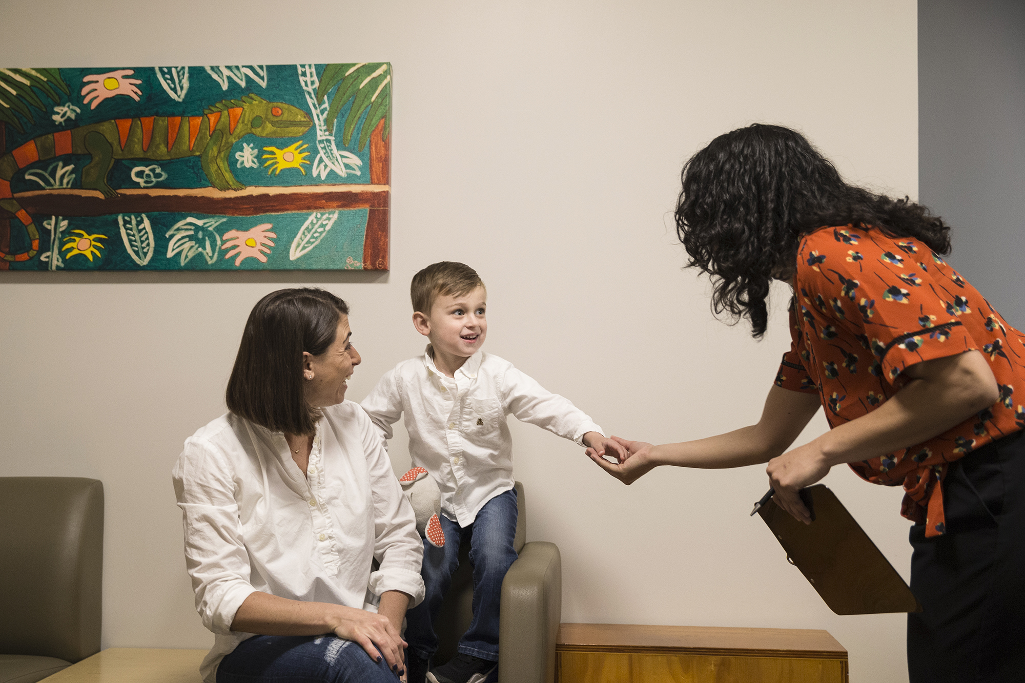 Photo: Owen and his mother sit on a chair in a doctor's office. A physician holding a clipboard is reaching out and grasping Owen's hand. He's looking at her with a pleasant expression on his face. His mother is smiling.