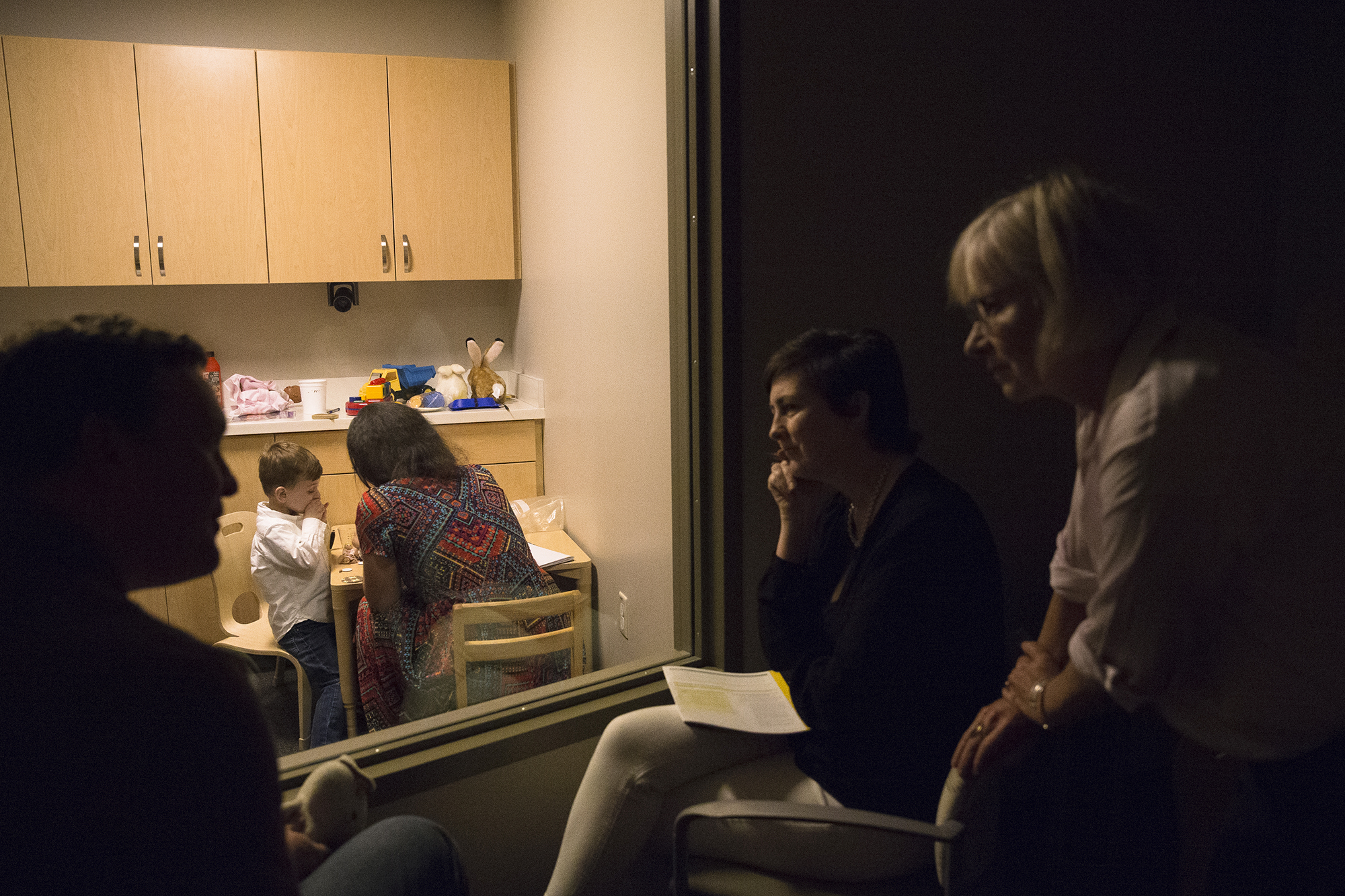 Owen and his mother sit at a table with a clinician while she tests his ability to follow instructions. Owen's father and two more clinicians watch through a window from another room.