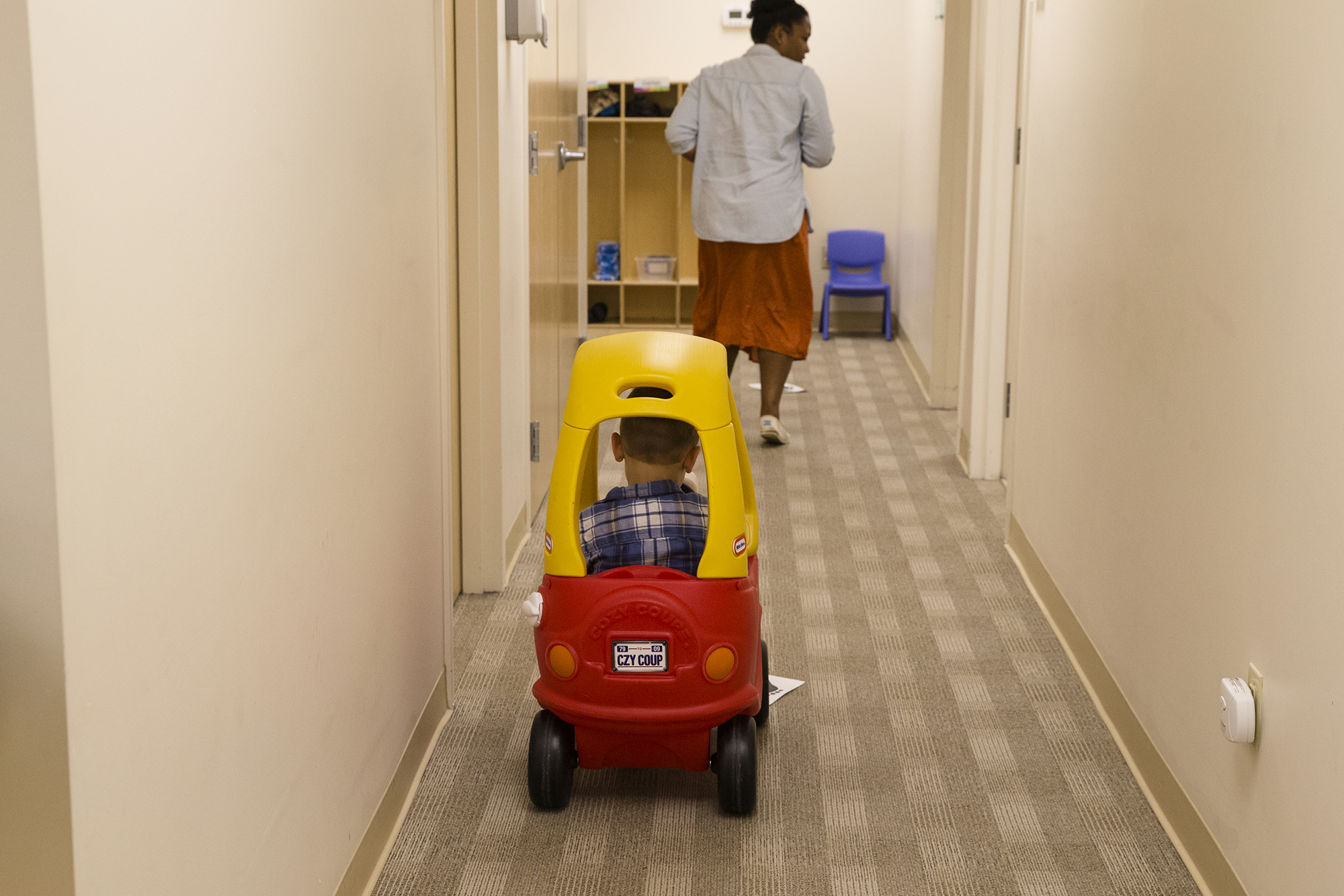 Photo: Owen plays in a yellow and red toy car, following a clinician down a carpeted hallway.