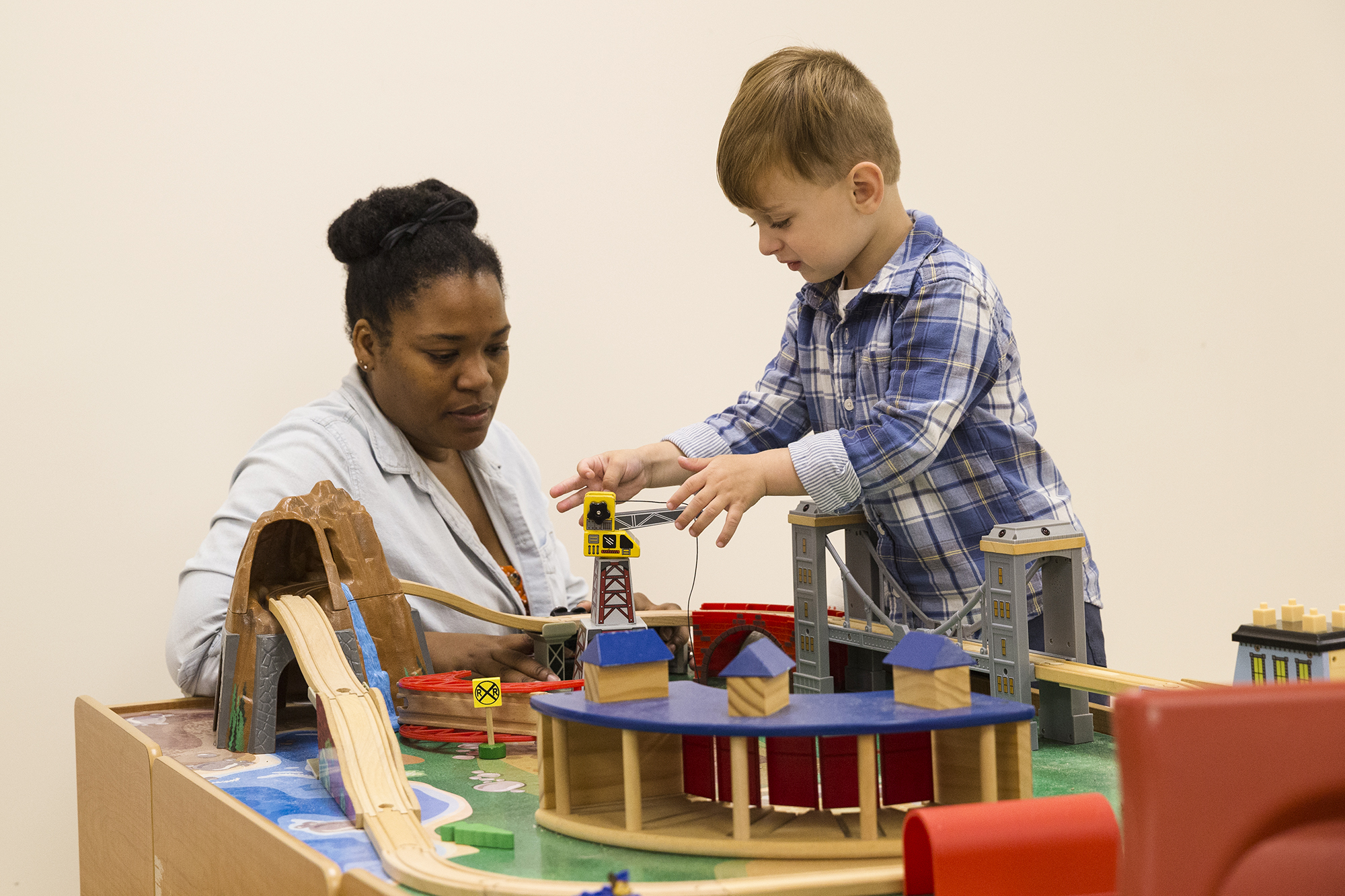 Owen stands at a play table, playing with toys, while a clinician observes.