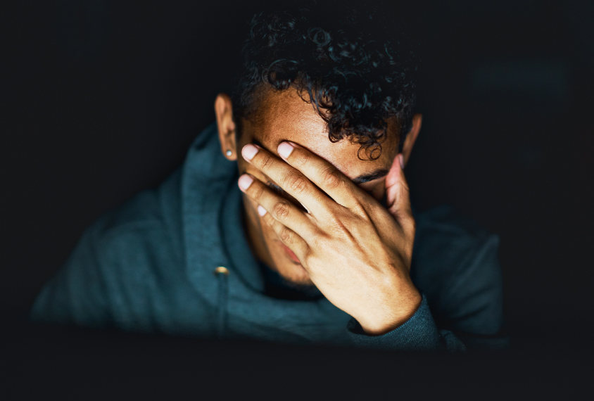 Young man holds his face in his hand, sitting in the dark.