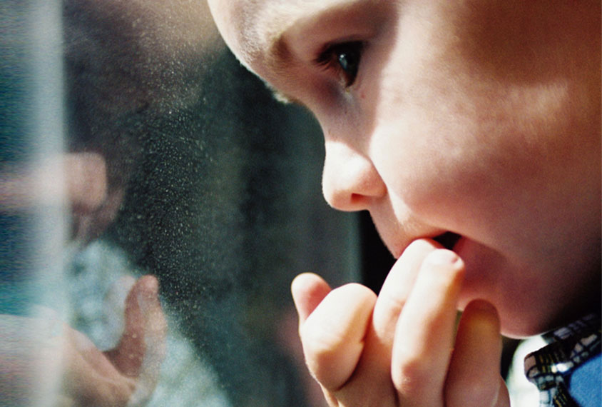 Young boy biting his fingernails, looking anxiously out the window.