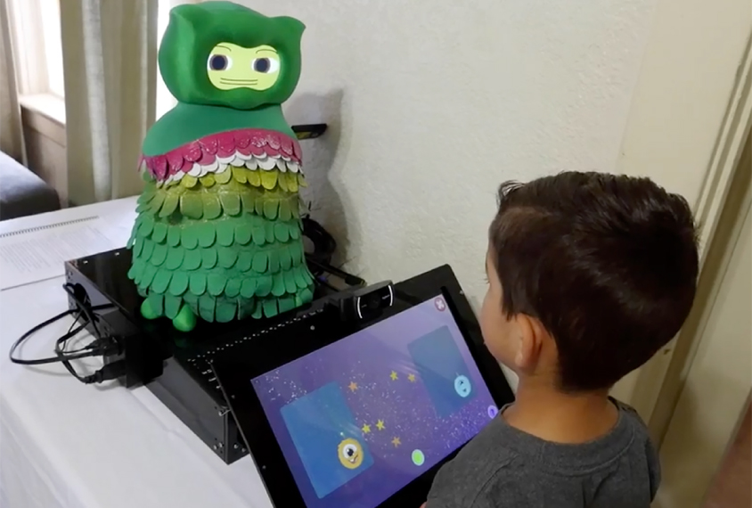 Young boy is interacting with a small rubbery green colored robot through a touch screen.