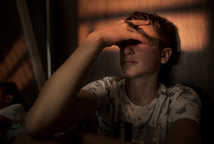 Teenage boy sitting alone, hand over his eyes, in the fading light.