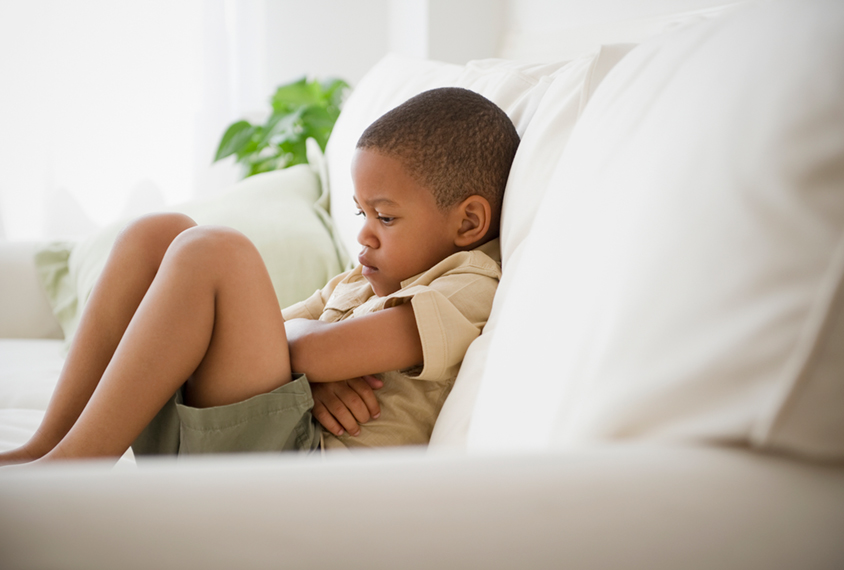 Young boy looking upset, sits on couch with crossed arms.
