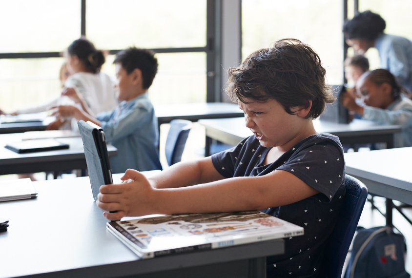 Boy in classroom looks frustrated as other kids interact and move around him.