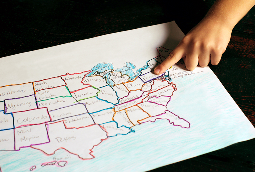 A child's hand points to a location on a hand drawn map of the United States in different crayon colors.