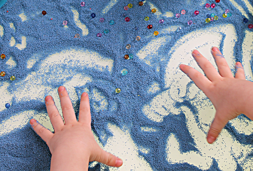 Child's hands playing on a white table with blue sand.