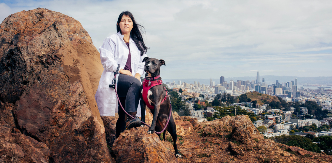 Portrait of Dr. Georgia Panagiotakos on a hill in San Francisco, with a view of the city behind her and her pet dog.