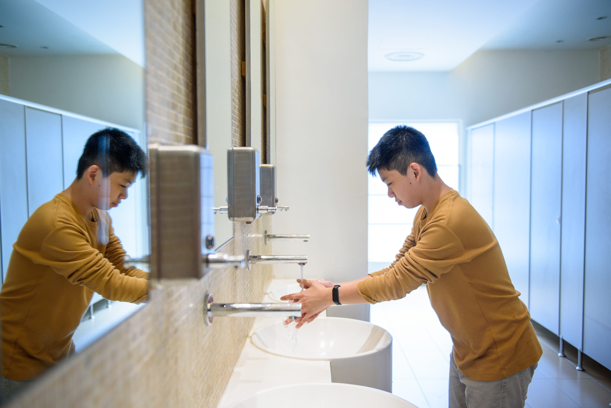 Photo: A boy in a yellow long-sleeve shirt washes his hands in a public restroom.