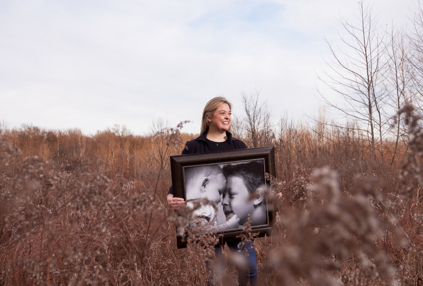 Portrait of Dr. Kristin Sohl in a field, with late afternoon light, holding a big black and white portrait of her children.