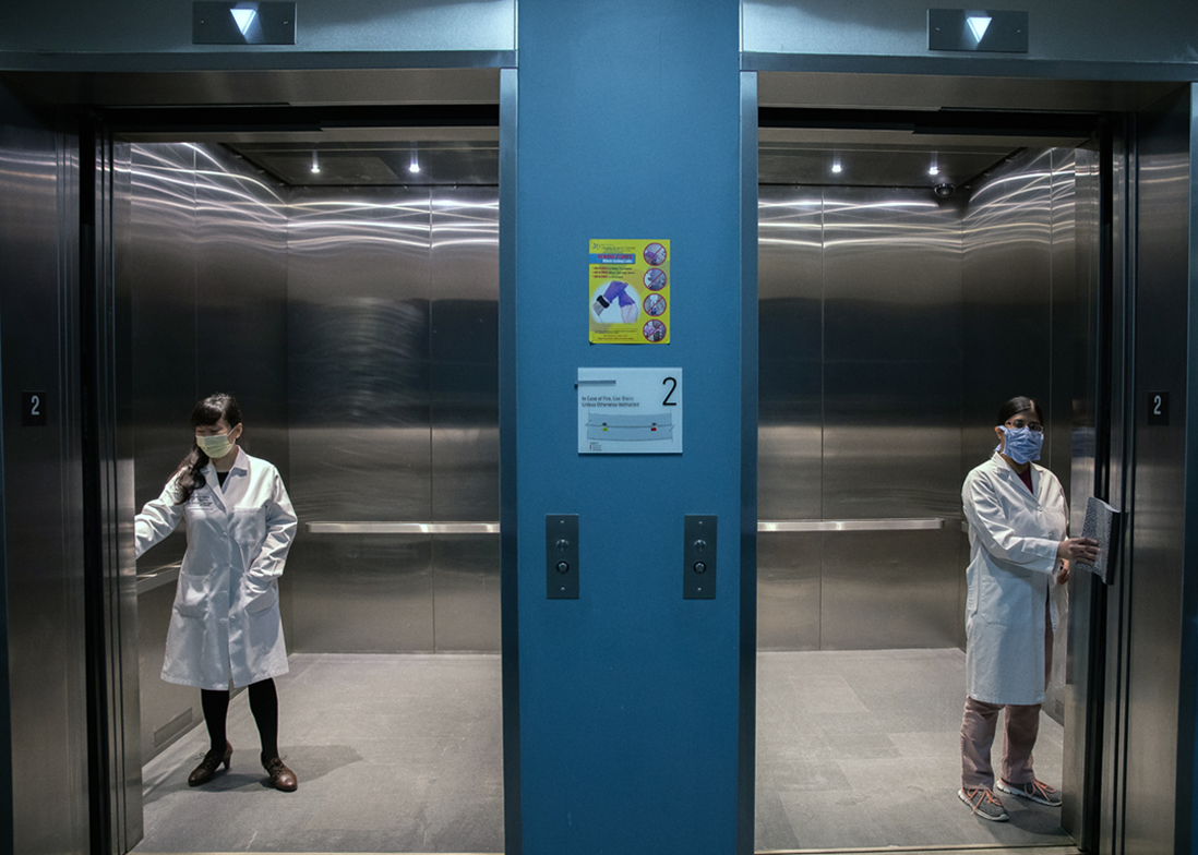 two researchers in two separate elevators, wearing COVID-19 protective gear