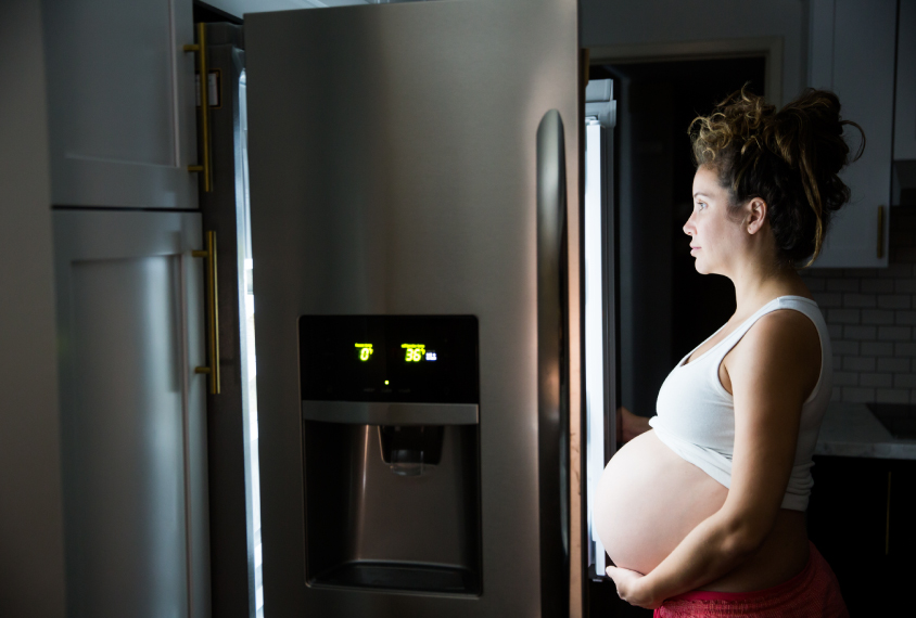 Pregnant woman standing in front of open refrigerator at night.