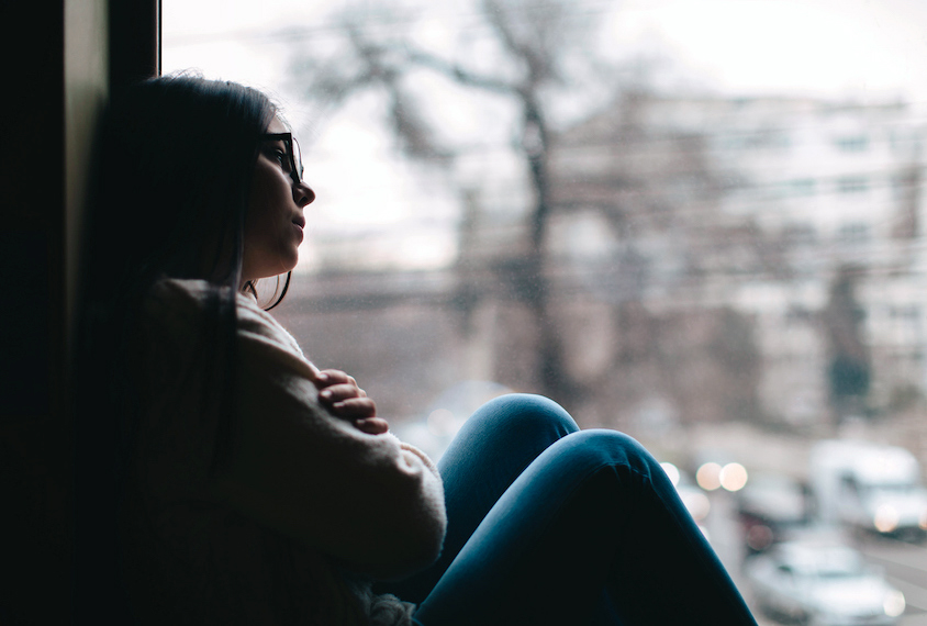 Young woman sitting alone at window in the shadows.