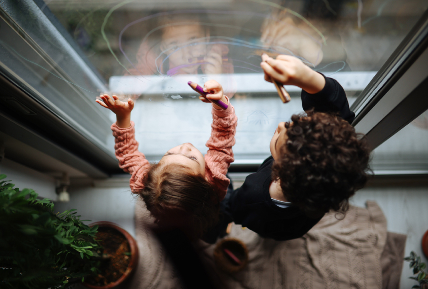 two toddlers, a boy and a girl, draw animals on a glass window in thin lines of blue, green and magenta.