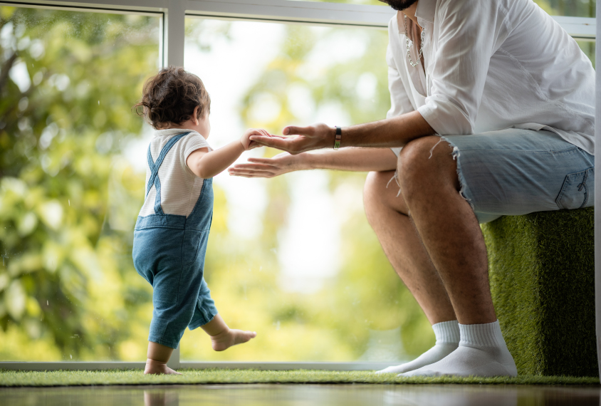 Toddler stepping towards parent, in front of window.