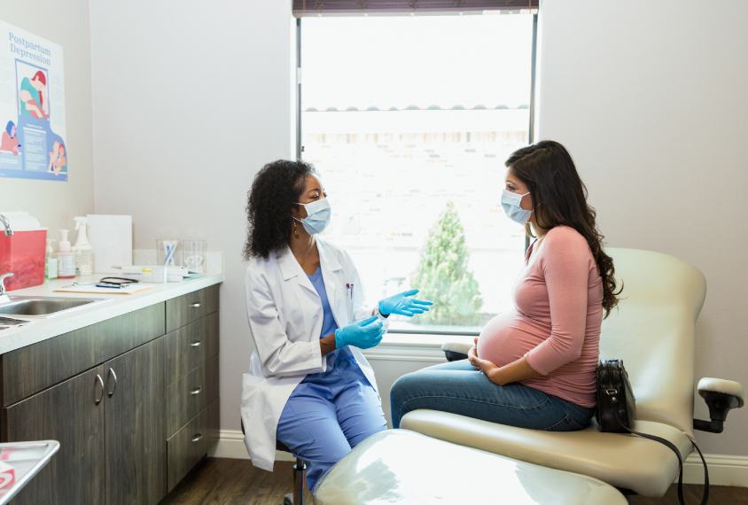 A pregnant woman and a doctor, both masked, have a conversation in an examination room.