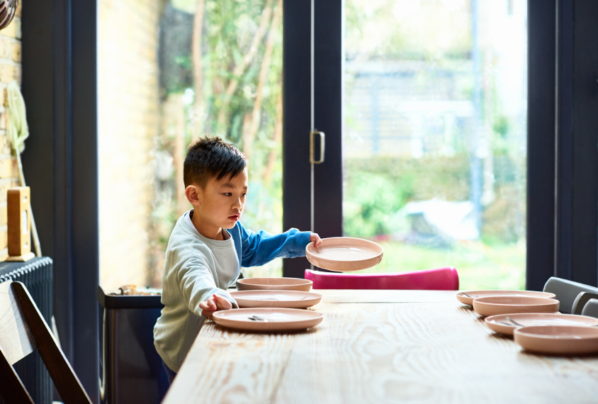 A young boy puts plates on a dining table.
