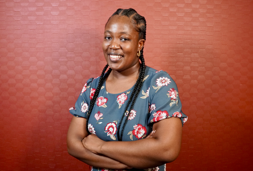 Oluwatobi Abubakare stands with their arms crossed against a light red background.