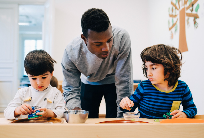 A teacher observes young students as they play with toys at a table.