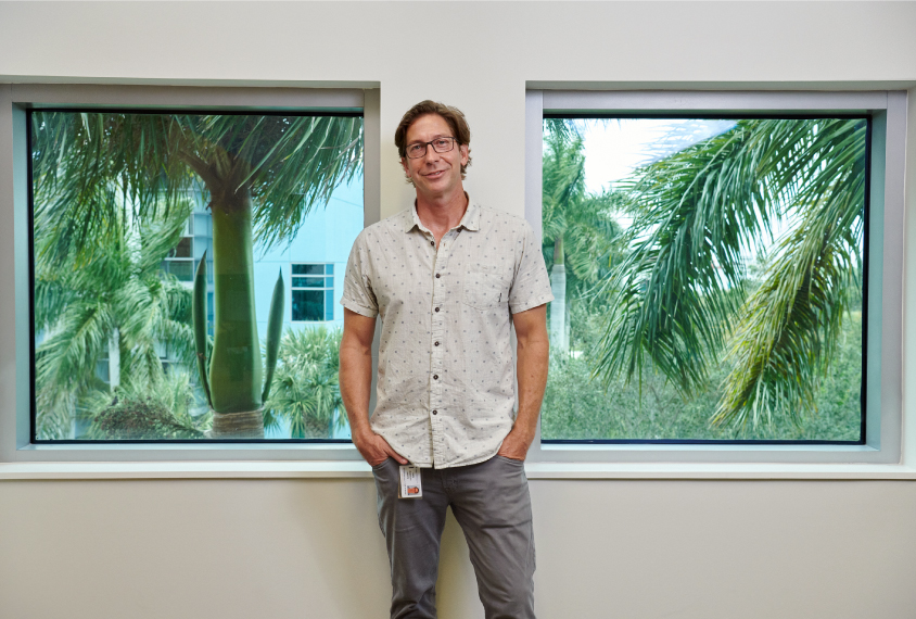 Gavin Rumbaugh stands with his hands in his pockets between two large windows with tropical trees and plants visible outside.